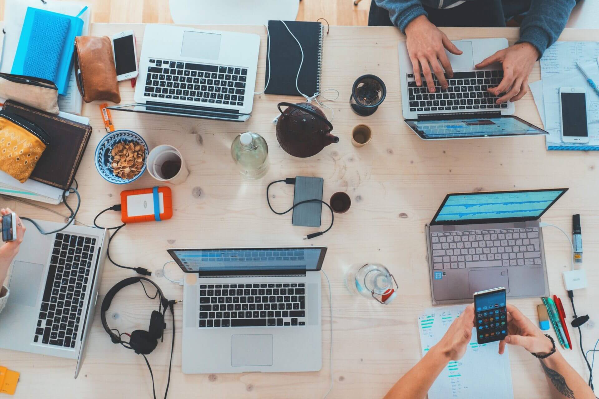 Laptops-smart-devices-headphones-hard-drives-hands-aerial Bird's-eye view of a work desk cropped in so you can see two sets of hands using laptops and smartphones, other electrical items are strewn across the table surface