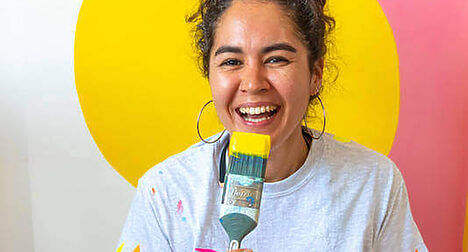 Portrait of medium-light skinned artist Eloisa Henderson-Figueroa, laughing with a paint brush and in front of a yellow and pink painted background