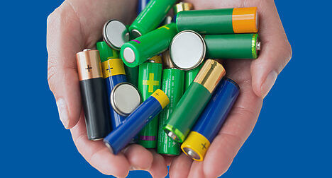 two light skinned hands holding a pile of household batteries against a blue background