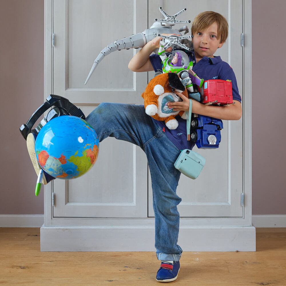 Boy balancing lots of toys header 2024 1000 px a light skinned boy holding lots of toys in his arms and balancing some on his legs as well