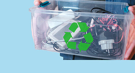 a light skinned person holding a see through plastic box full of electrical goods that has the green recycling symbol on it