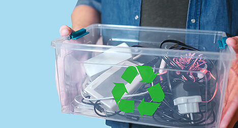 light skinned person holding a see-through plastic box of old cables, with a green recycling symbol on the front