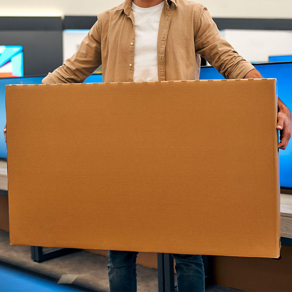 Man with TV in a cardboard box TVs shutterstock 2108275586 1000 px light skinned man holding a very large cardboard box with a TV inside.
