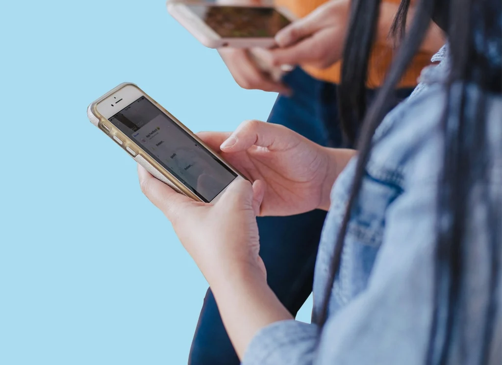 troubleshooting-with-phone copy Medium light skinned young person using a smart phone, with someone else using a phone in the background.