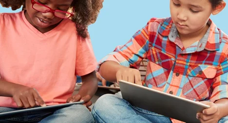 Two children, one dark skinned wearing glasses, one medium-light skinned, sitting cross legged using tablets against a bright blue background.