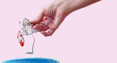 a pale skinned hand holding a white cable above an open blue bin against a pink background