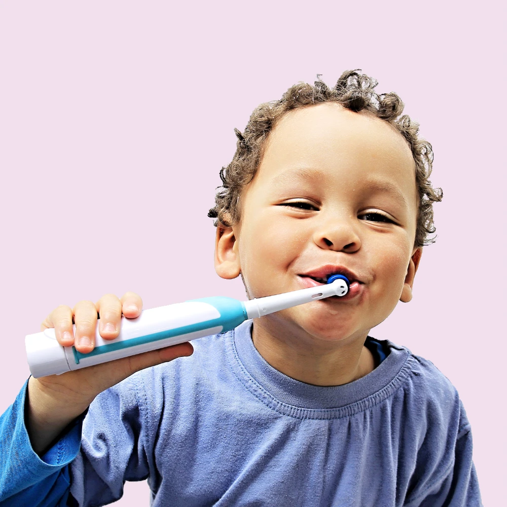 Electric toothbrush boy shutterstock_1193664295 1000 px medium skin tone boy brushing teeth with an electric toothbrush and smiling, against pink background