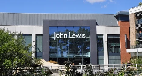 Entrance of a John Lewis store with shiny glass windows reflecting the blue sky and trees surrounding the building.